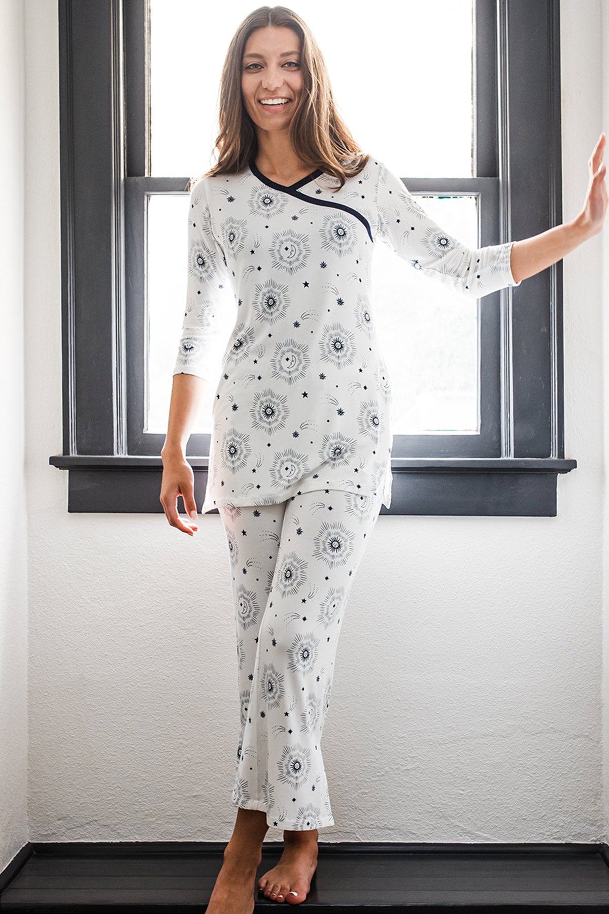 A woman standing in front of a window and leaning against a wall while smiling, wearing Yala Haley Crossover Front 3/4 Sleeve Bamboo Pajama Set in Celestial Print