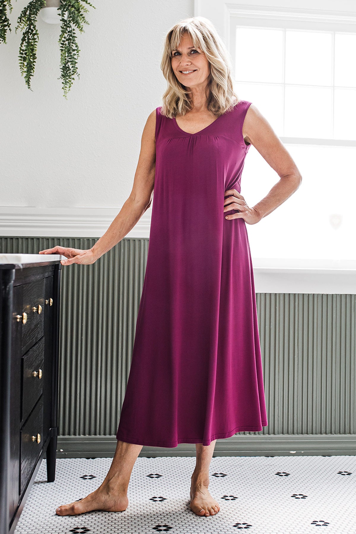 A woman standing next to cabinet and smiling, one hand on top of the cabinet and the other resting on her hip, wearing Yala Molly Sleeveless Bamboo Nightgown in Boysenberry