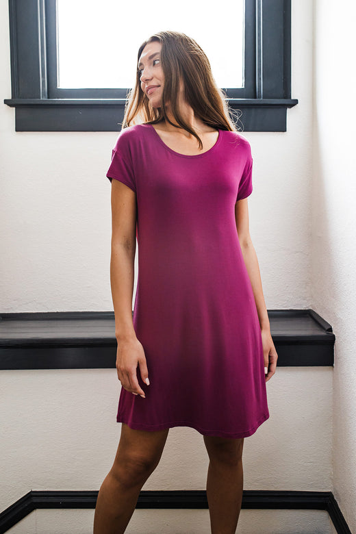 A woman standing in front of a window while looking up and to the side, wearing Yala Penelope Cap Sleeve Oversized Bamboo Nightshirt in Boysenberry