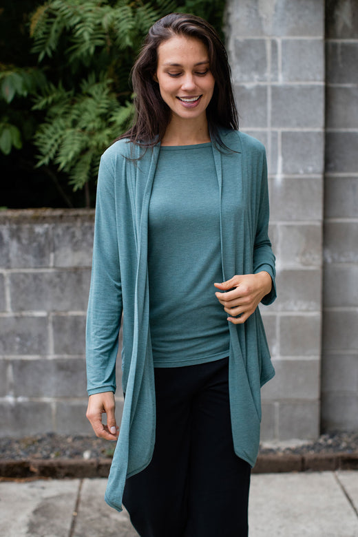 A woman standing and smiling, looking down with one hand holding her jacket hem, wearing Yala Sophie Bamboo Cardigan Wrap in Teal Melange