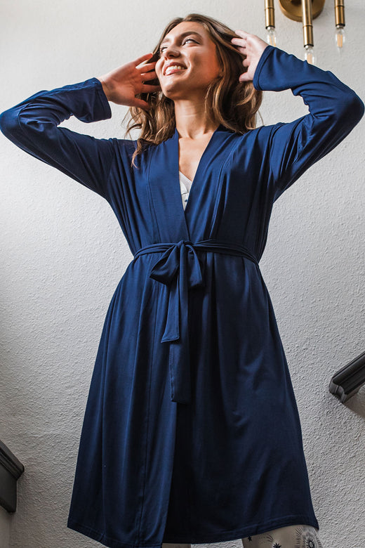 A woman standing and looking up and off to the side with her hands in her hair, wearing Yala Serenity Long Sleeve Short Belted Bamboo Robe in Navy