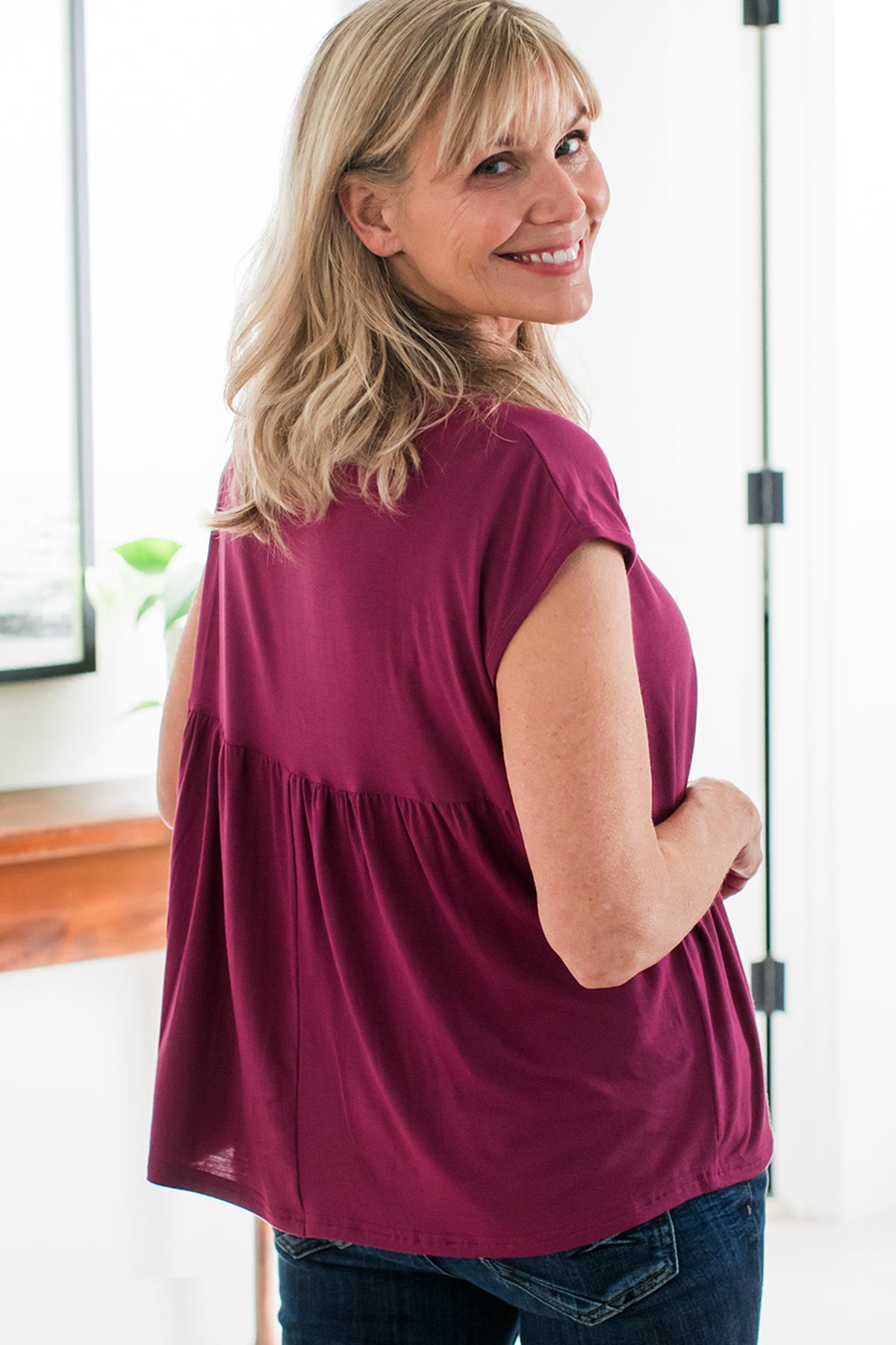 A woman standing with her back to the camera, smiling and looking back over her shoulder, wearing Yala Opal Swing Bamboo Top in Boysenberry