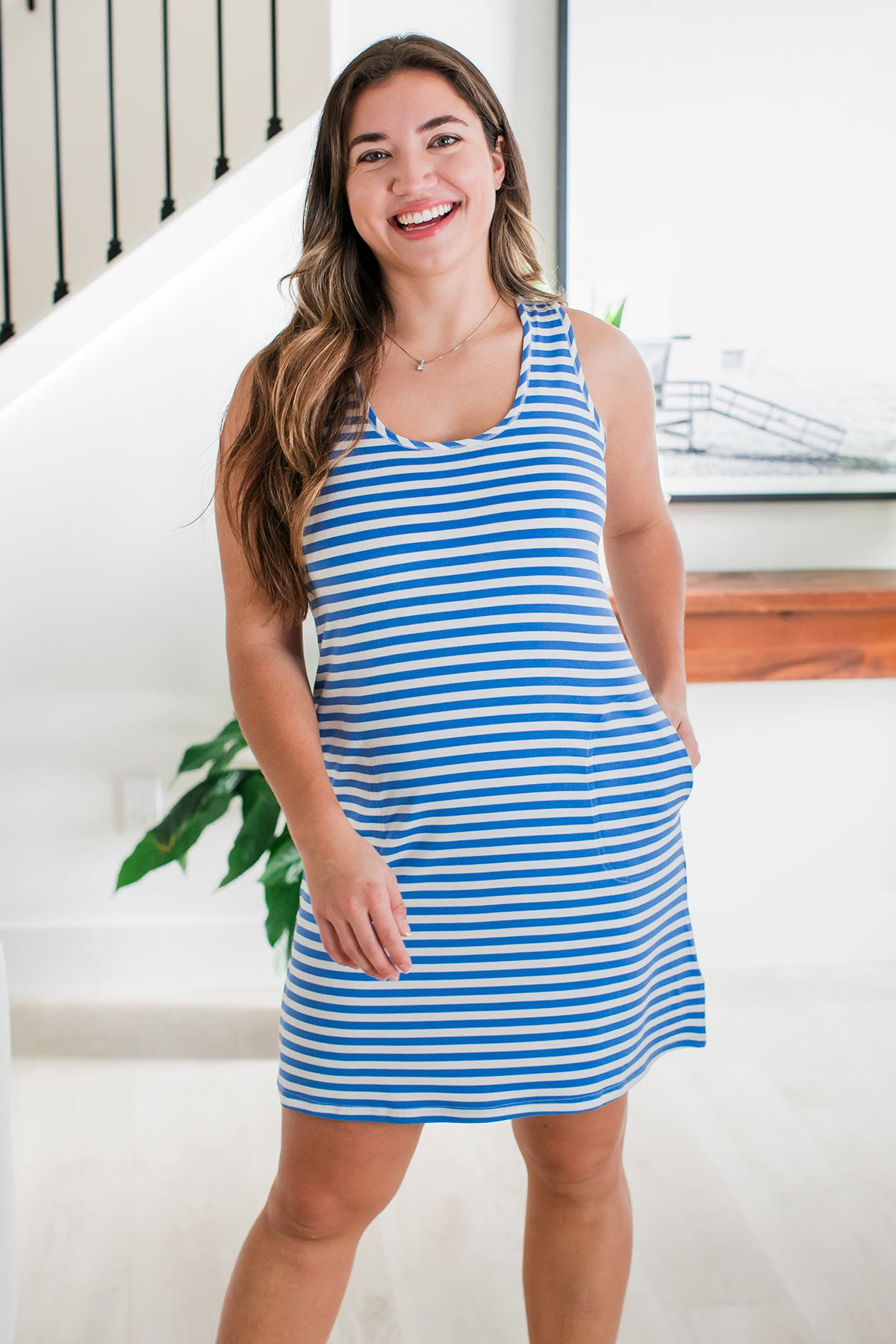 A woman standing with a smile on her face and one hand in her pocket, wearing Yala Riley Racerback Two Pocket Bamboo Shift Dress in French Blue Natural Stripe