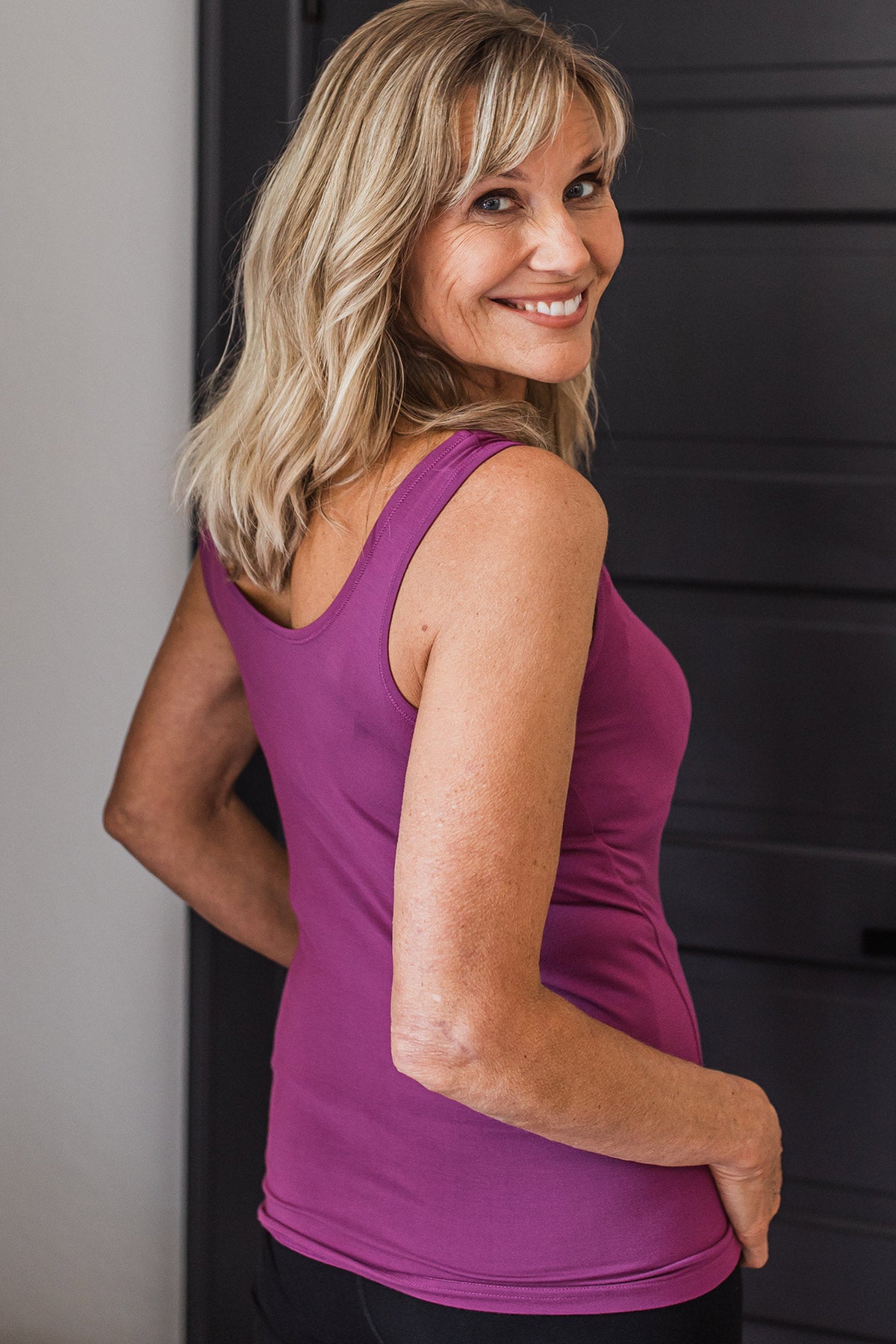 A woman standing facing away from the camera, looking back over her shoulder and smiling with her hands on her hips, wearing Yala Zia Layering Bamboo Tank Top in Boysenberry