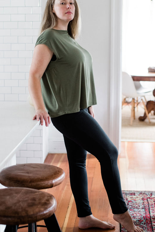 A woman standing and leaning back against a counter with one leg outstretched, wearing Yala Beckett High-Waisted Bamboo and Organic Cotton Skinny Pants in Black