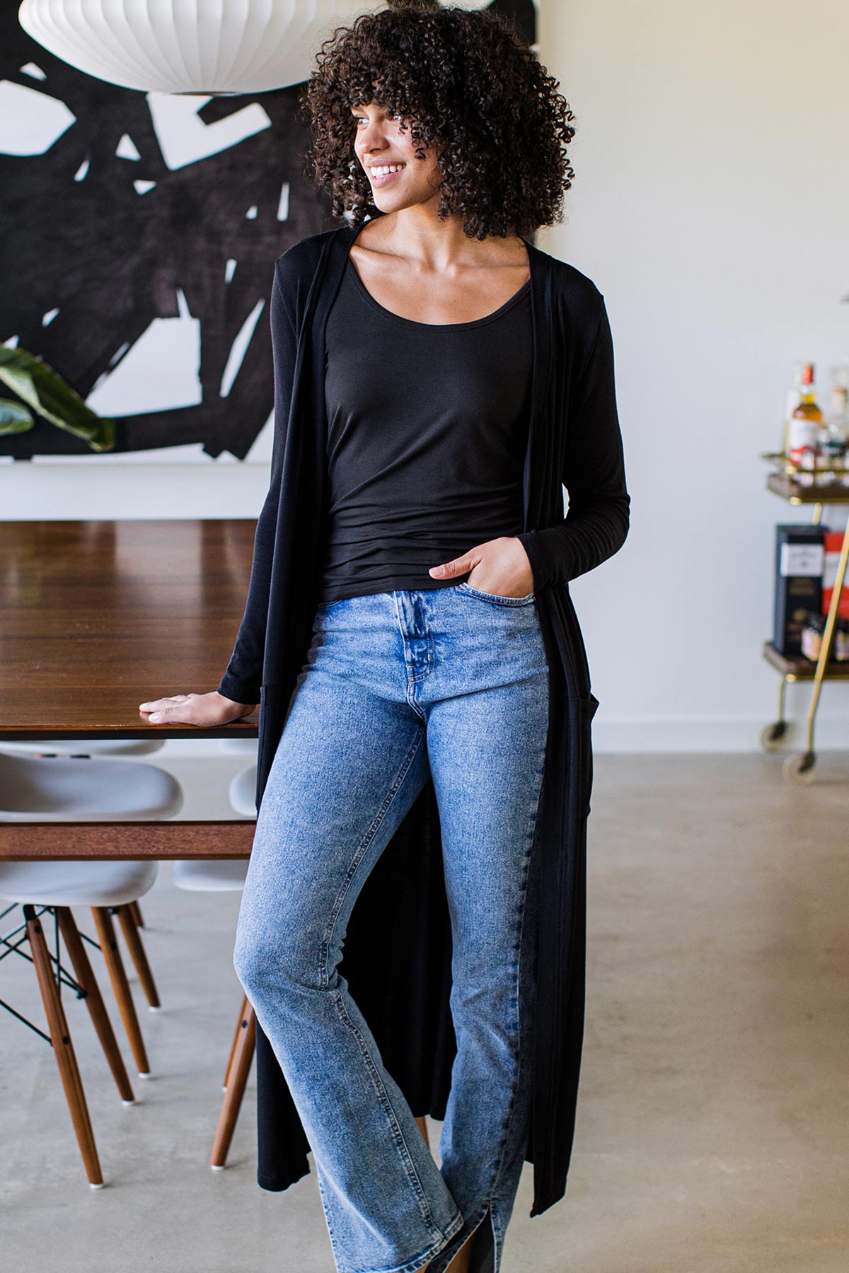 A womand standing and smiling while looking to the side with one hand in her belt line, wearing Yala Brooke Cardigan Duster Bamboo Sweater in Black