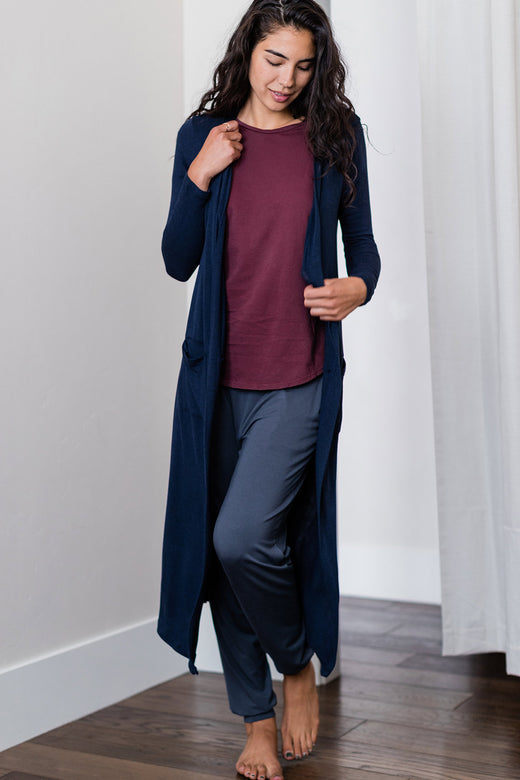 A woman standing with one knee bent while looking downwards, wearing Yala Brooke Cardigan Duster Bamboo Sweater in Navy
