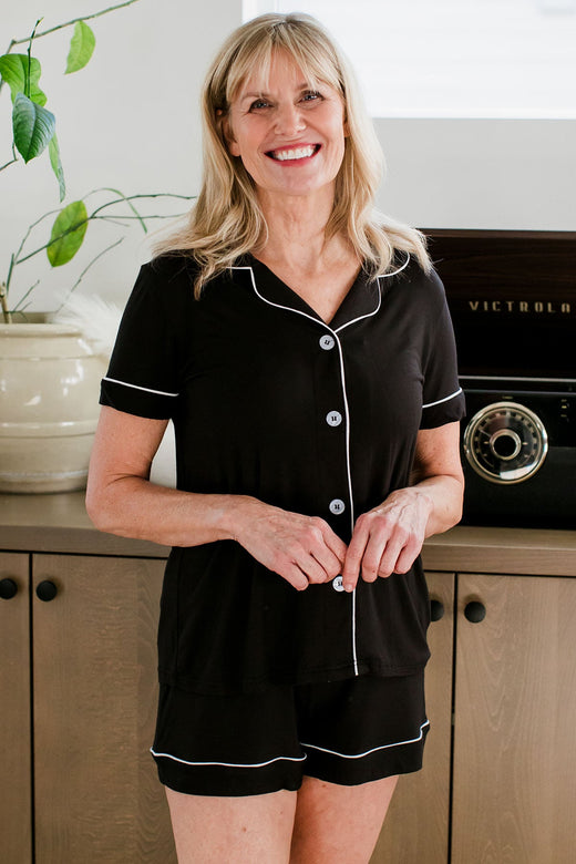 A woman standing with both hands clasped in front of her and a smile on her face, wearing Emma Classic Button Front Shorts Bamboo Pajama Set in Black