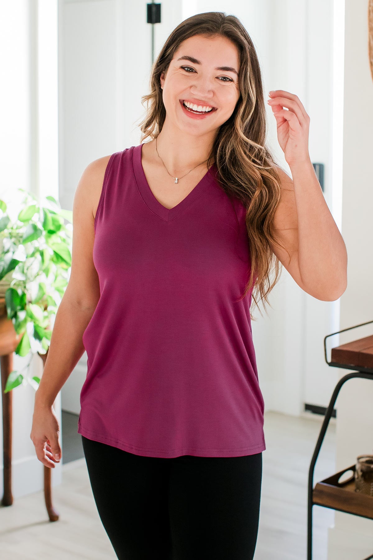 A woman standing with on hand raised towards her hair and a smile on her face, wearing Ginger V-Neck Racerback Bamboo Tank Top in Boysenberry