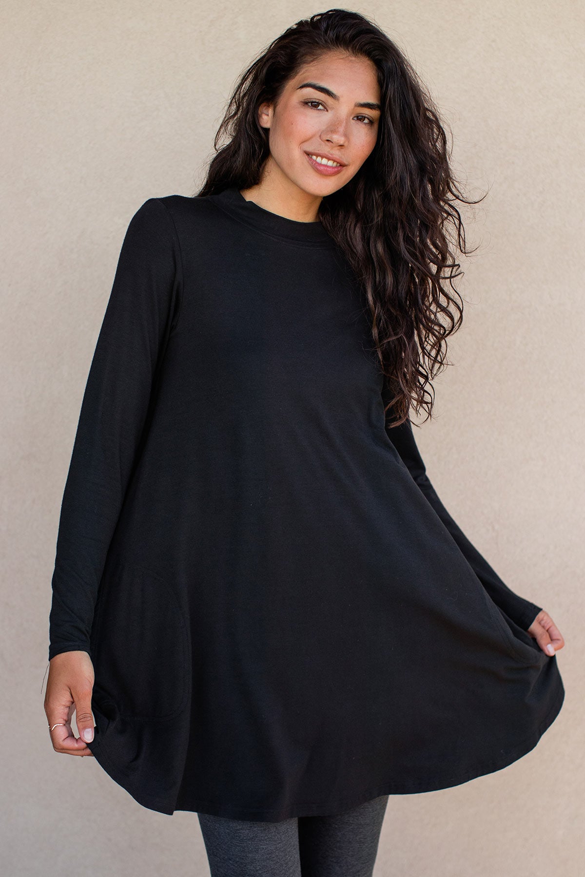 A woman standing and smiling while holding out the edges of her dress, wearing Yala Cadence Mock Neck Bamboo Swing Dress in Black