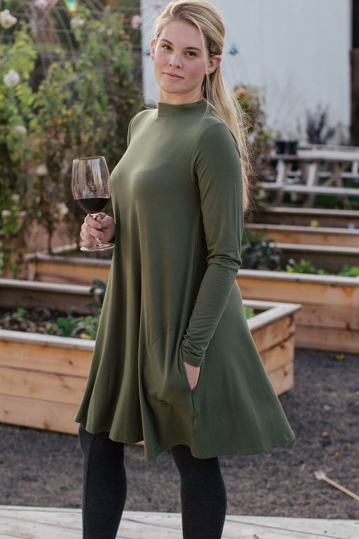 A woman standing facing to the side and looking at the camera with one hand in her pocket and the other holding a glass of wine, wearing Yala Cadence Mock Neck Bamboo Swing Dress in Moss