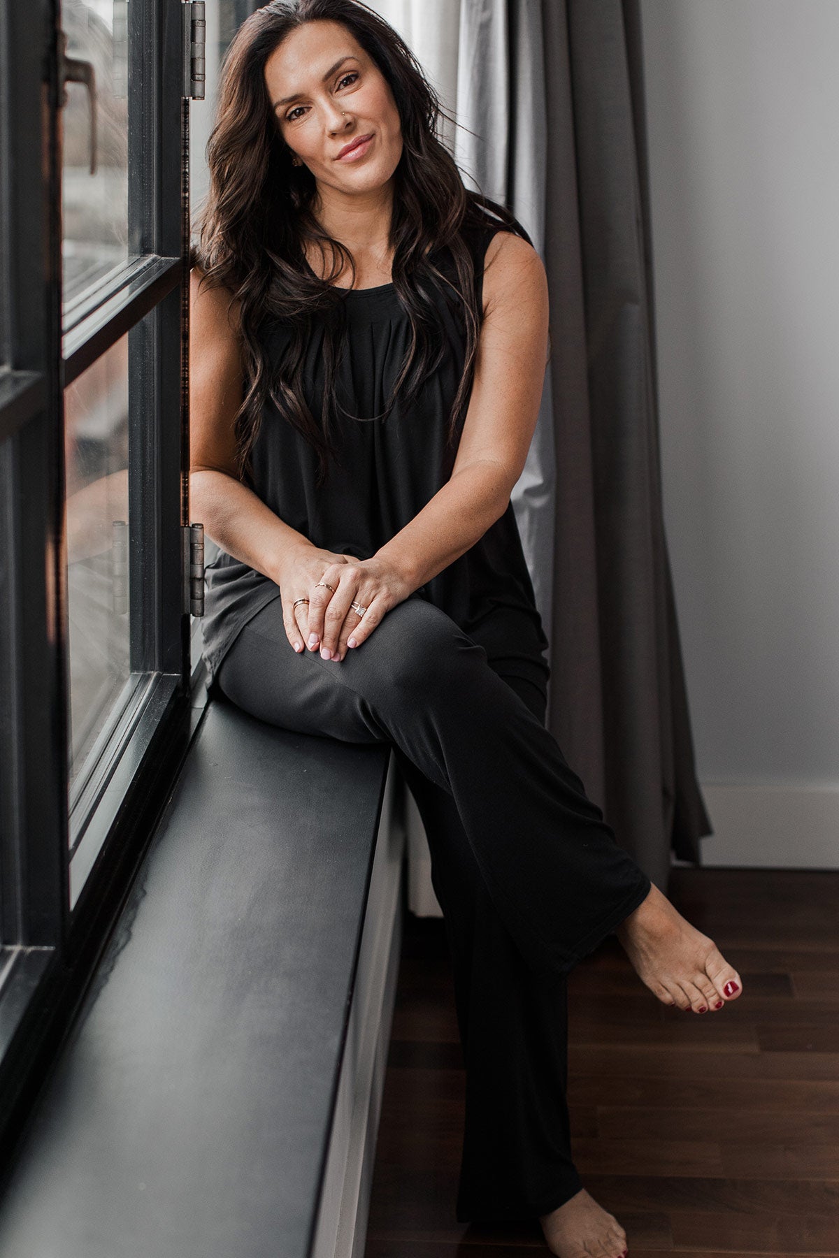 A woman sitting on a windowsill with her knees crossed and her hands on her thigh, wearing Yala Delia Gathered Tank Bamboo Pajama Set in Black