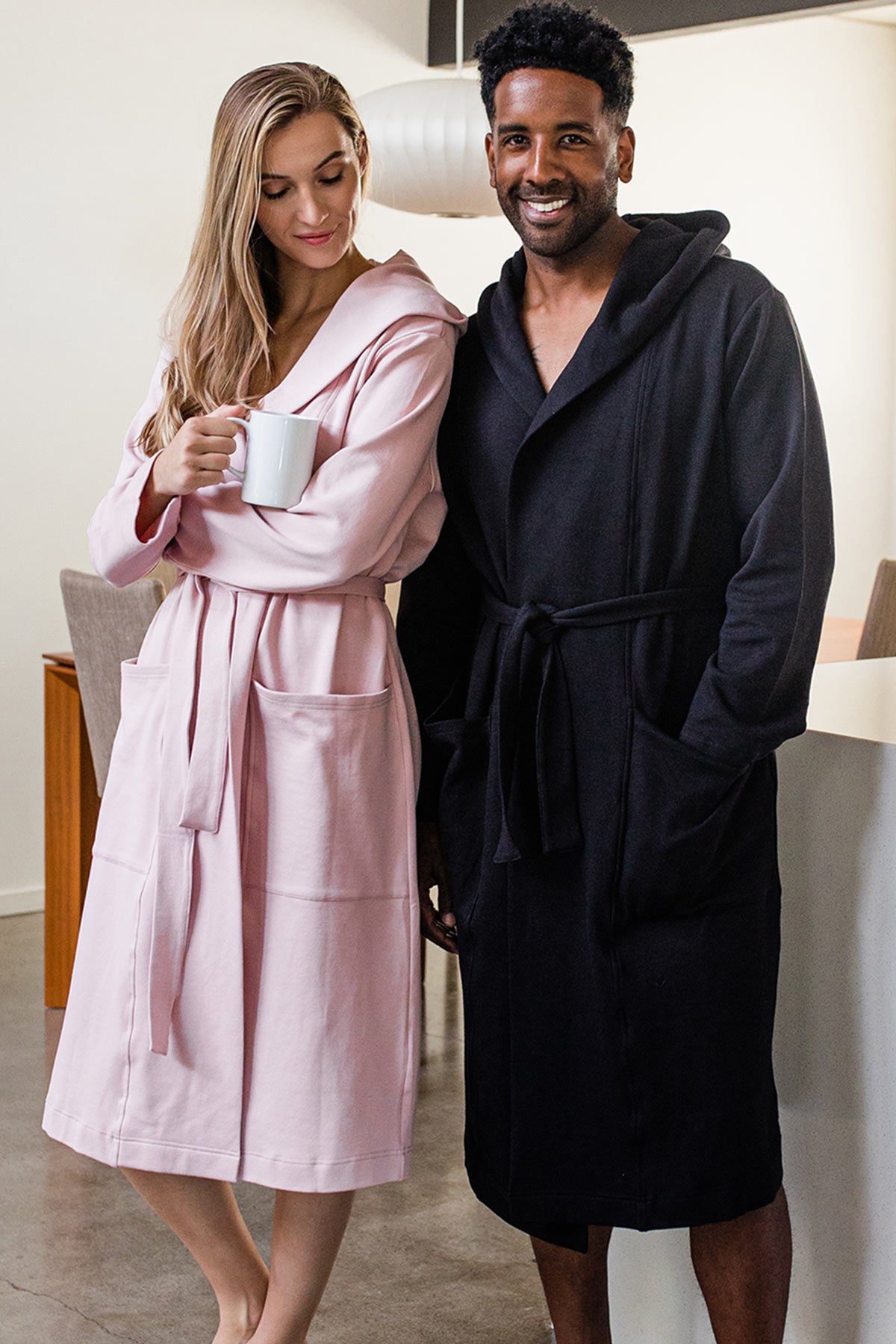 A man and woman standing together, the woman leaning against the man, both wearing Yala Elliot Bamboo and Organic Cotton Hodded Robe in Black and Lotus Pink