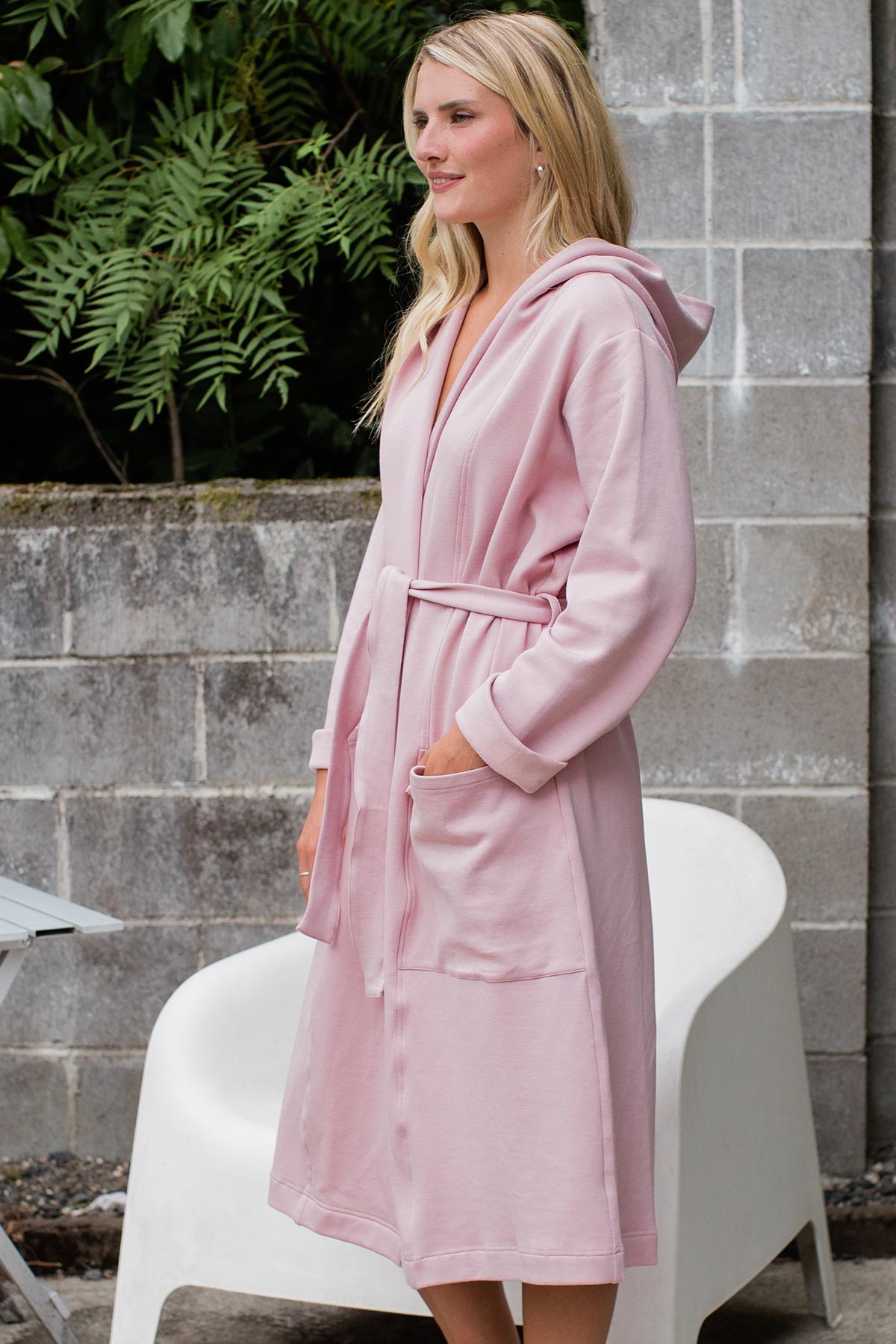 A woman standing facing to the side with one hand in her robe pocket, wearing Yala Elliot Bamboo and Organic Cotton Sweatshirt Hooded Robe in Lotus Pink