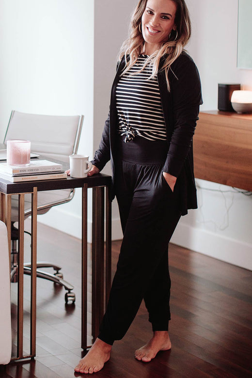 A woman standing with one foot forward and one hand holding a cup on a tabletop, wearing Yala Geena Lightweight Bamboo Jogger Lounge Pants in Black