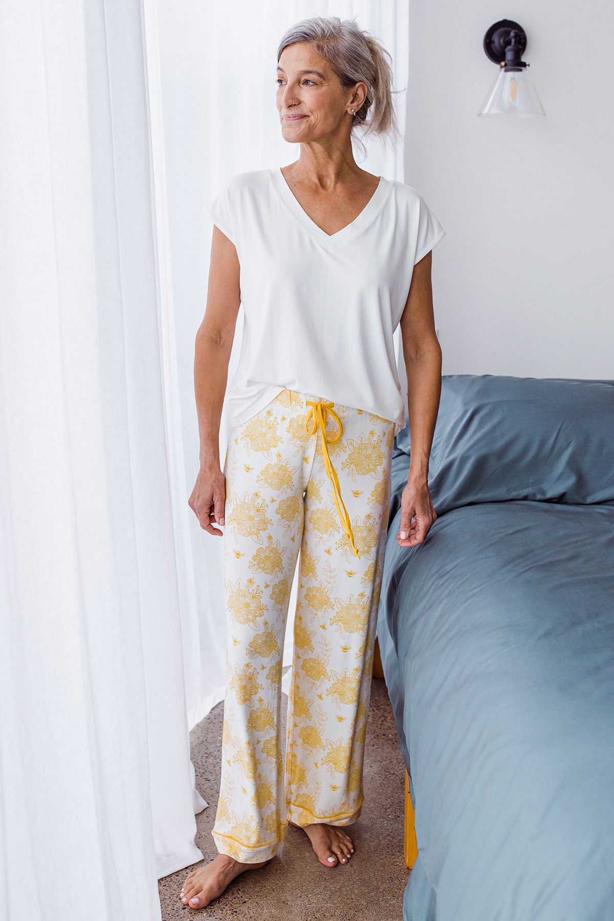 Woman standing and looking out a window, wearing Yala Gillian Piped Bamboo Pajama Pants in Honeybee