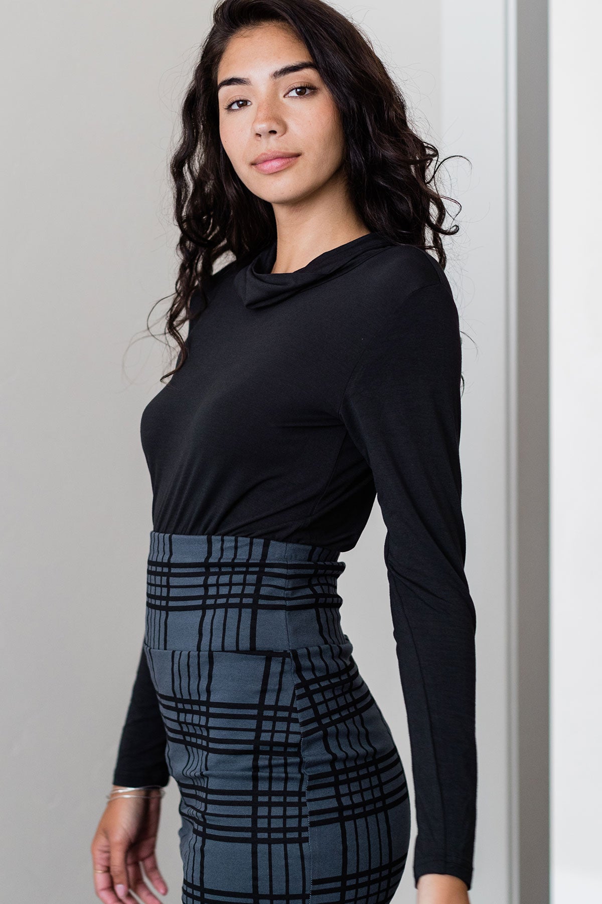 A woman standing facing to the side and looking towards the camera, wearing Yala Harper Cowl Neck Long Sleeve Bamboo Shirt in Black