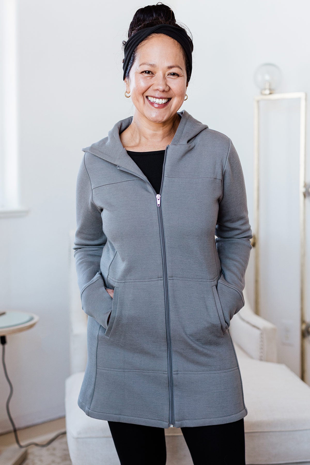 A woman standing and smiling with both hands in her jacket pockets, wearing Yala Jemma Zip-Up Long Bamboo and Organic Cotton Sweatshirt Hooded Jacket in Space Grey