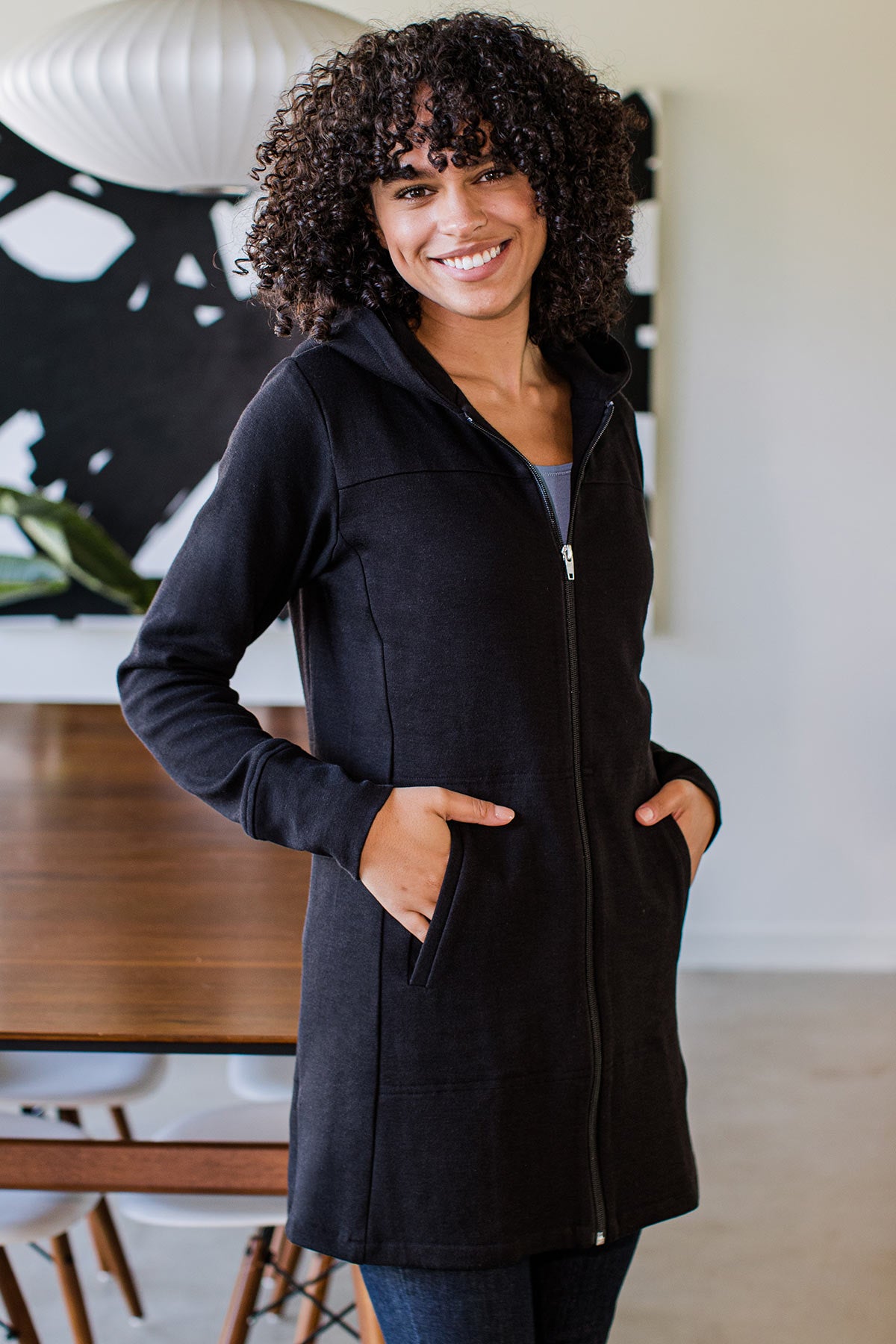 A woman smiling and standing sideways with both hands in her jacket pockets, wearing Yala Jemma Zip-Up Long Bamboo and Organic Cotton Sweatshirt Hooded Jacket in Black
