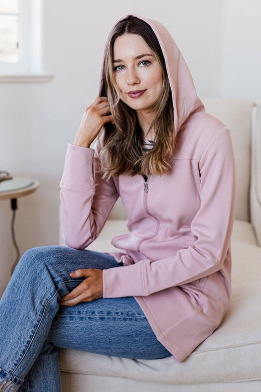 A woman sitting on a chaise lounge with her knees crossed, wearing Yala Jemma Zip-Up Long Bamboo and Organic Cotton Sweatshirt Hooded Jacket in Lotus Pink