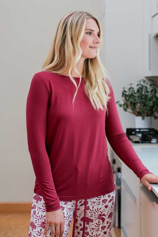 A woman standing and smiling while looking sideways out of a window, wearing Yala Jena Long Sleeve Bamboo Tee Shirt in Rosewood