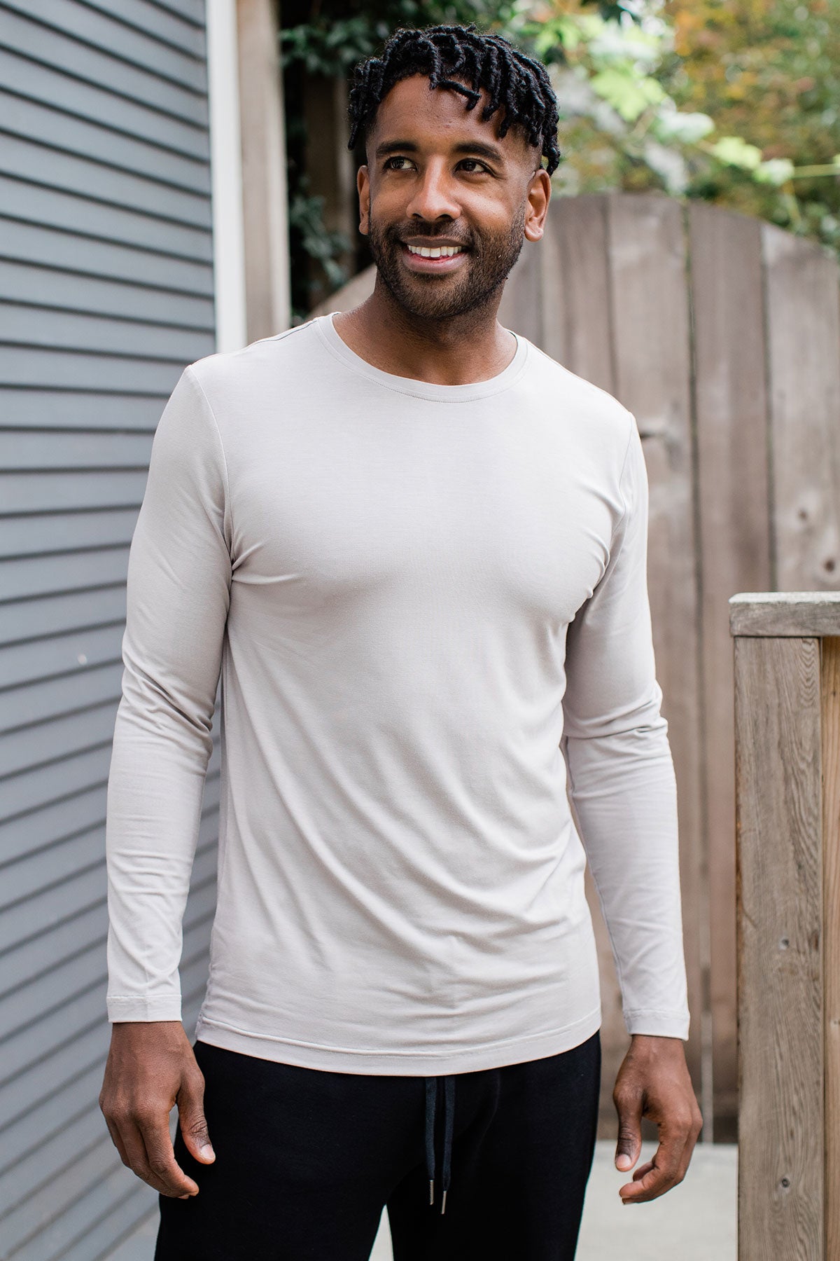 A man standing and smiling while looking to the side, wearing Yala Jonah Men's Long Sleeve Bamboo Crew Tee Shirt in Ash
