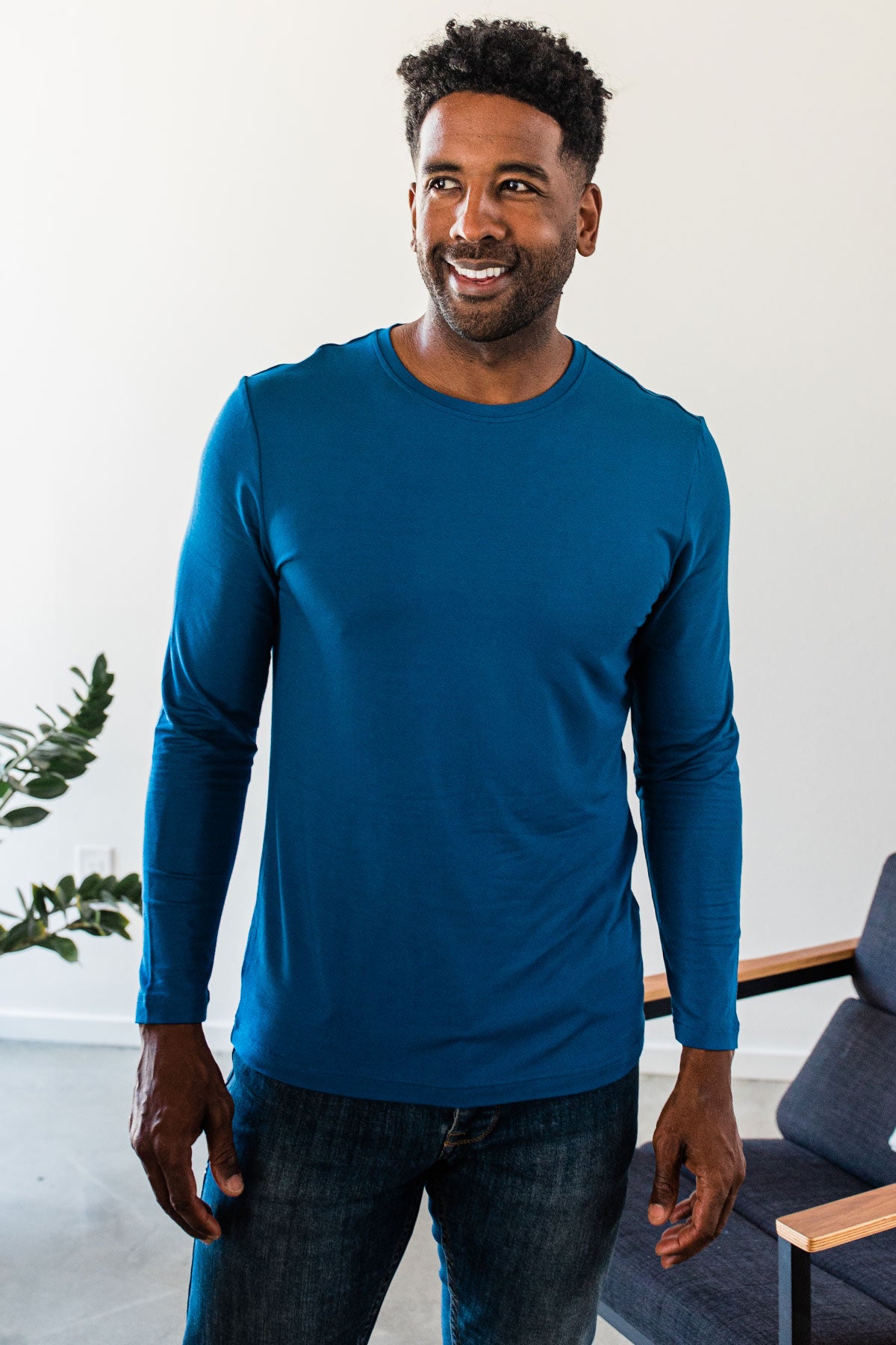 A man standing and smiling while looking to the side, wearing Yala Jonah Long Sleeve Bamboo Crew Tee Shirt in Lapis