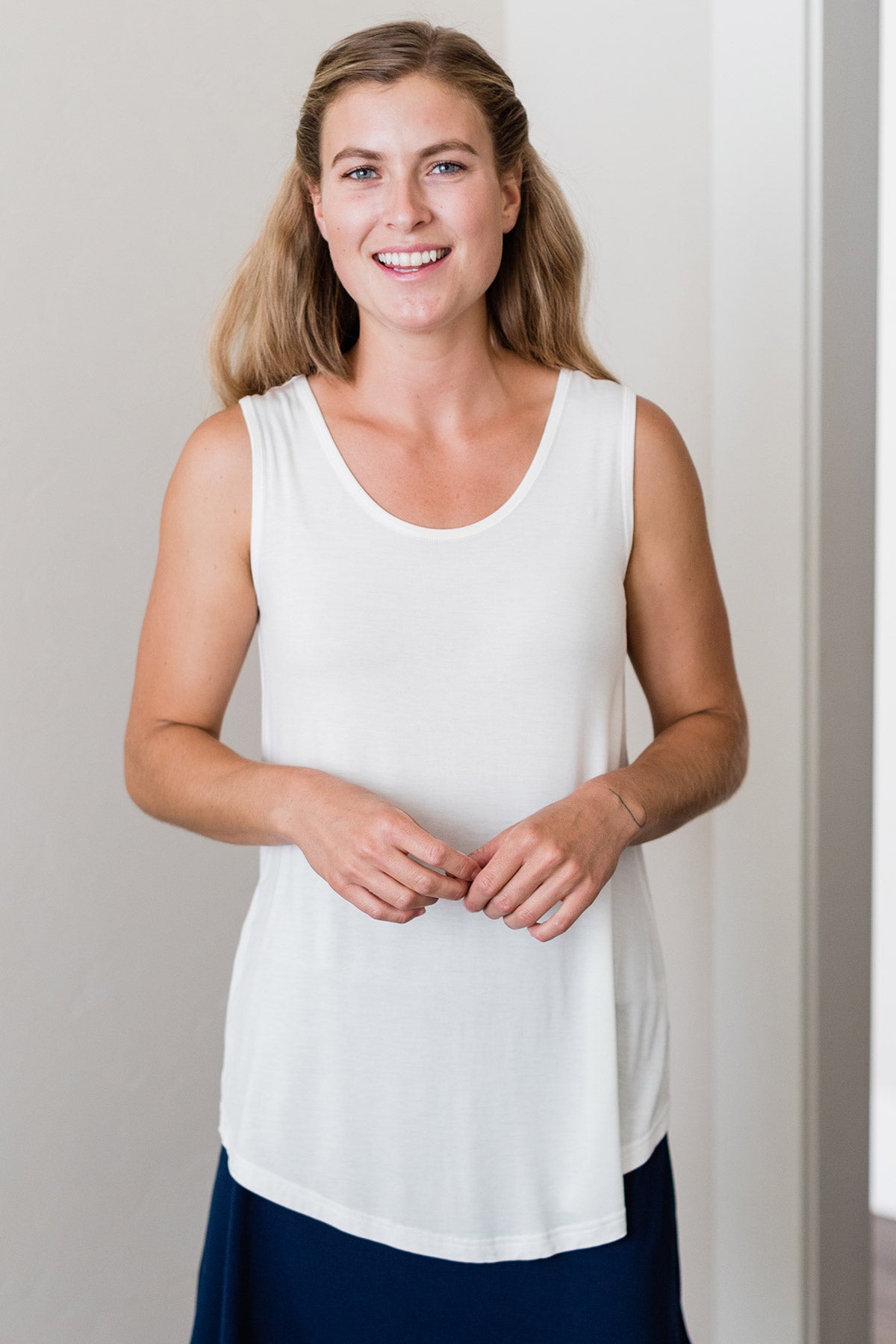 A woman standing and smiling with both hands held in front of her, wearing Yala Justine Bamboo Tunic Tank Top in Natural