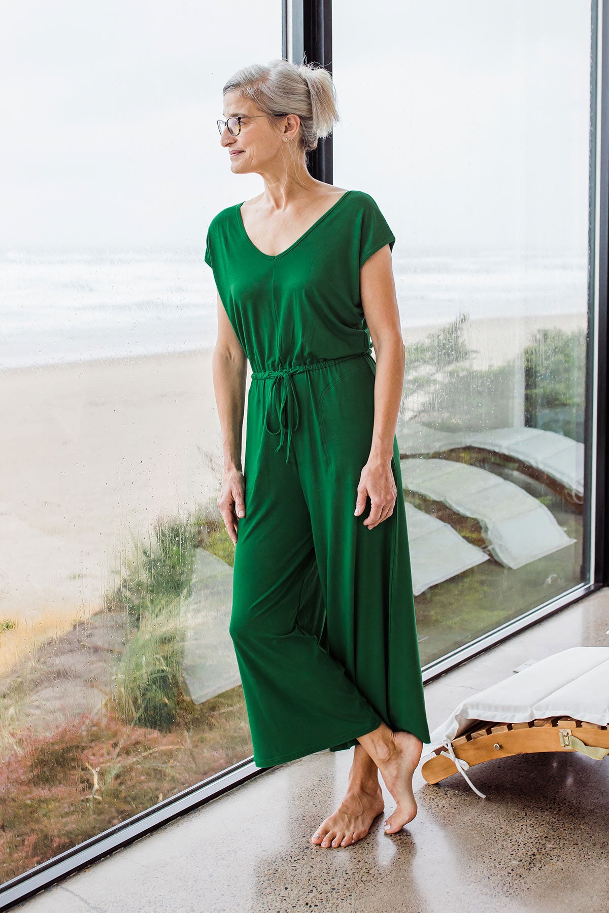 Woman standing and leaning against a window, wearing Yala Kiova V-Neck Bamboo Jumpsuit in Emerald