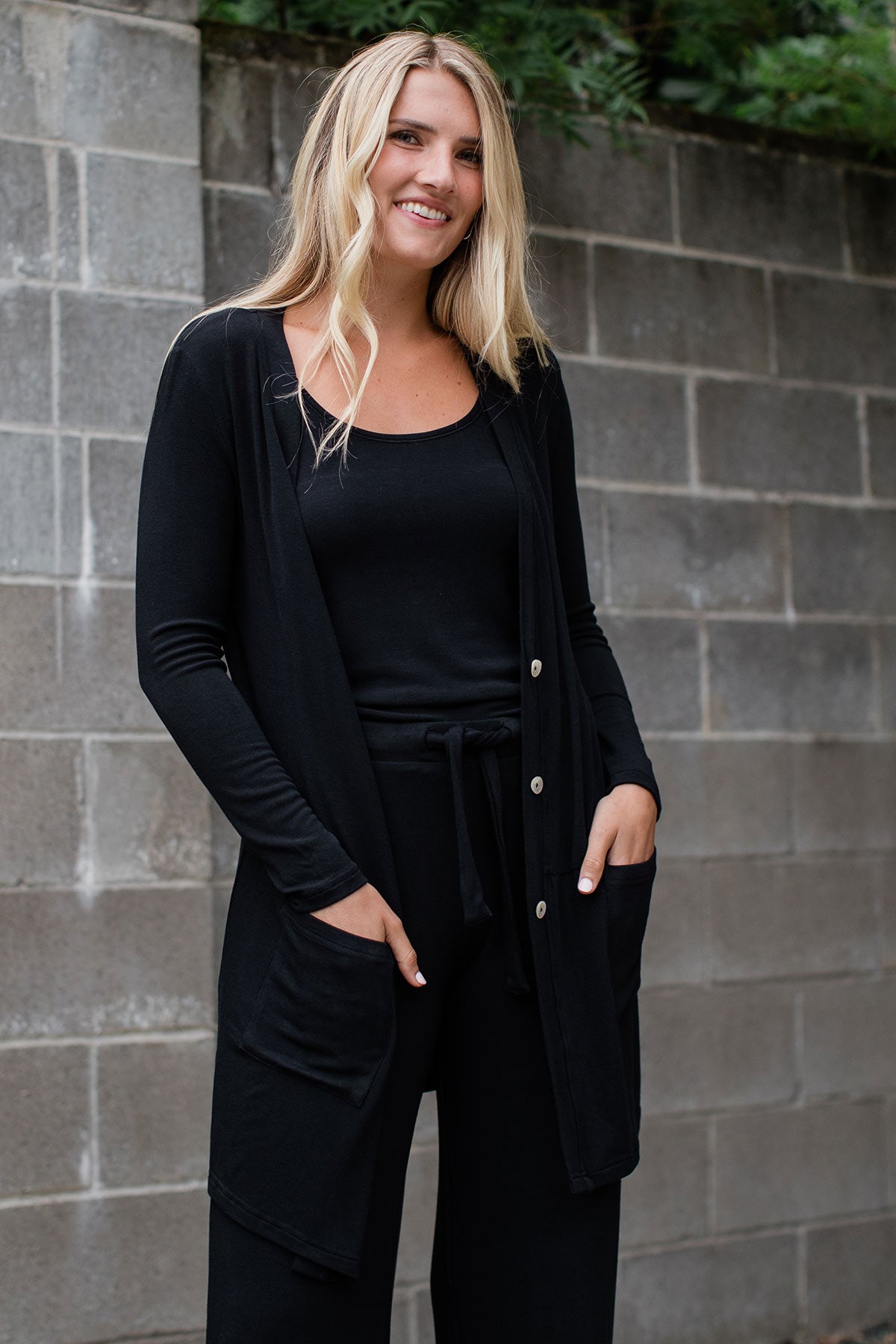 A woman standing and smiling with both hands in her pants pockets, wearing Yala Leslie Long Sleeve Oversize Bamboo Cardigan in Black