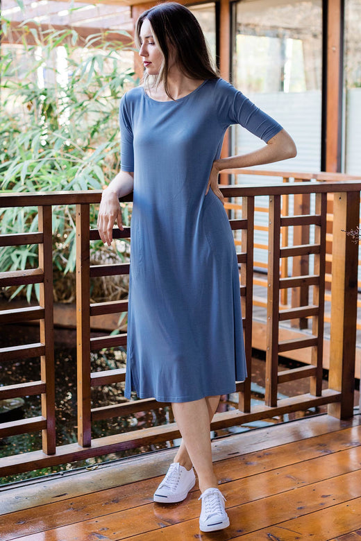 A woman standing with one arm resting on railing and legs crossed, wearing Yala Maraya Midi Elbow Sleeve Bamboo Dress in Denim