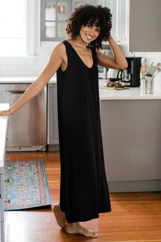 A woman standing sideways and smiling with one arm stretched behind her to rest on a countertop, wearing Yala Molly Sleeveless Bamboo Nightgown in Black