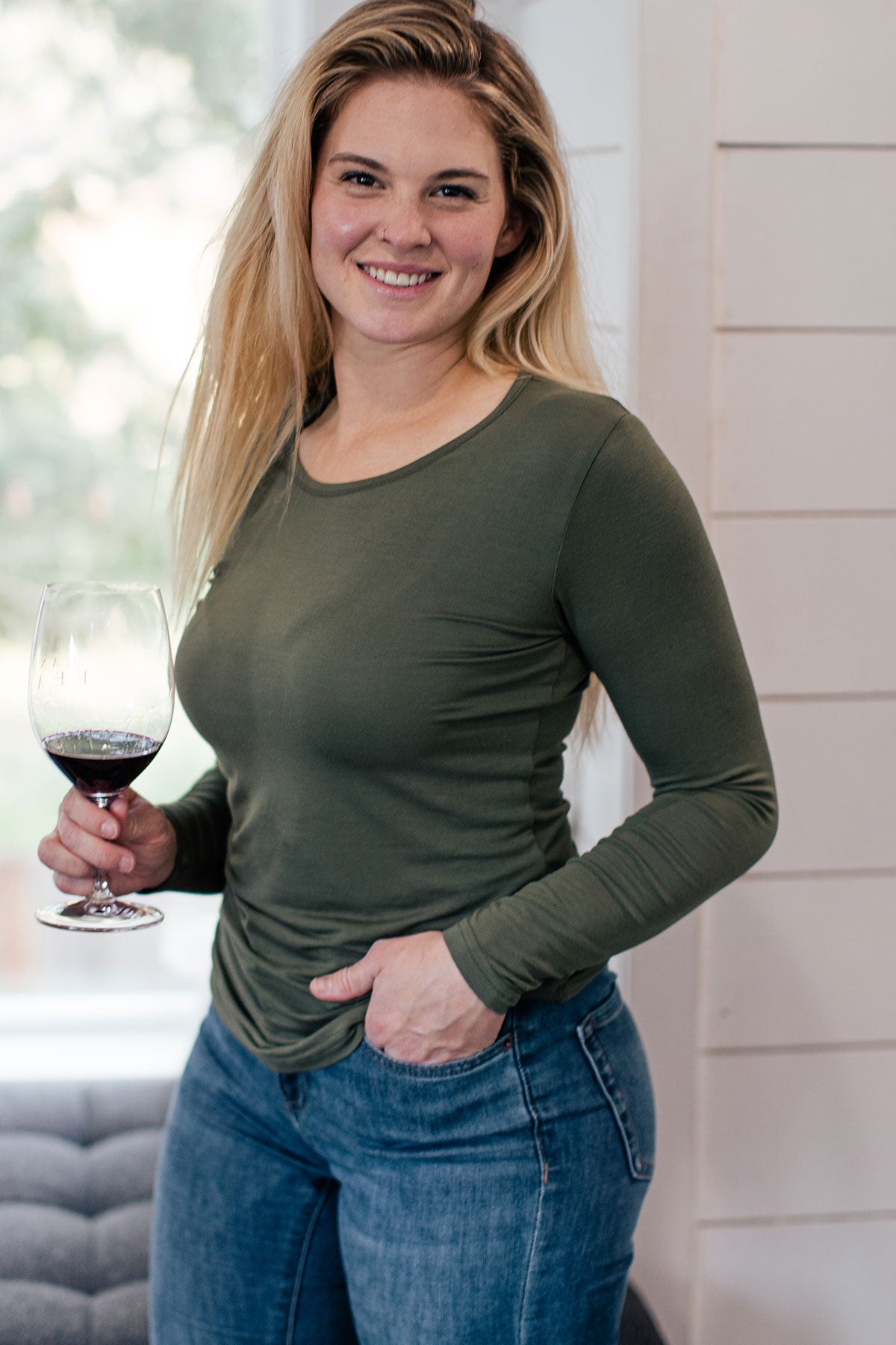 A woman standing and smiling with one hand in her pocket and the other holding a glass of wine, wearing Yala Natalie Long Sleeve Bamboo Crew Tee Shirt in Moss