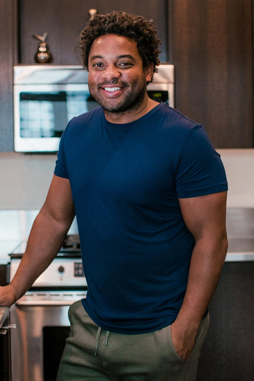 A man standing and smiling with on hand resting on a countertop and the other in his pocket, wearing Yala Nathan Men's Short Sleeve Bamboo Crew Tee Shirt in Navy