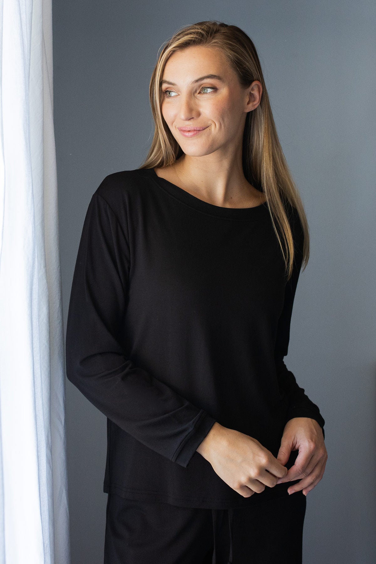 A womand standing a gazing out of a window smiling, wearing Yala Navarre Bamboo Sailor Tee in Black