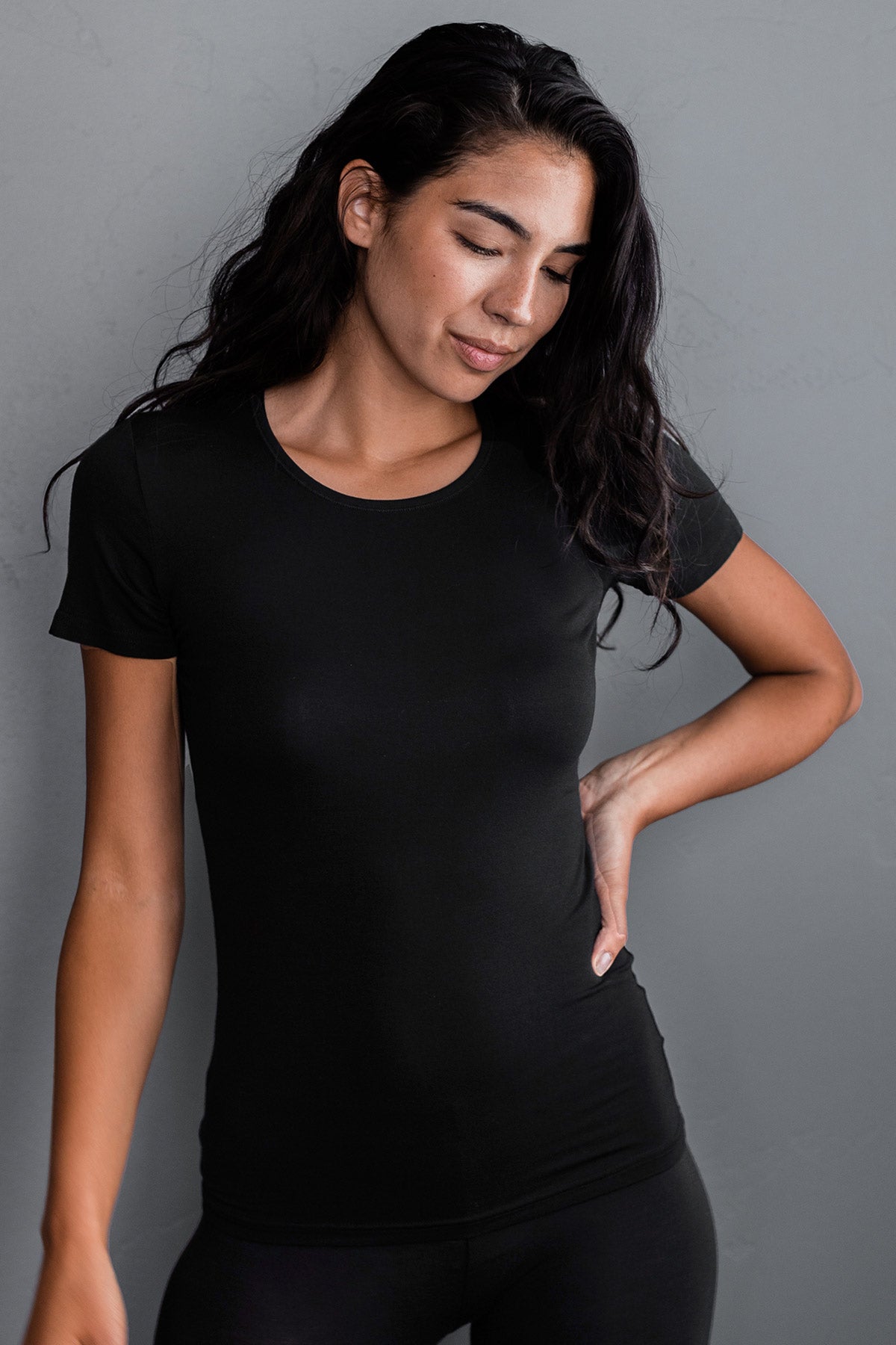 A woman standing and looking downward with one hand on her hip, wearing Yala Nicole Short Sleeve Fitted Bamboo Tee Shirt in Black