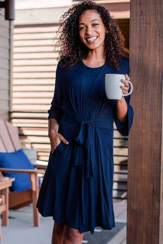 A woman standing and leaning against a wall with a cup of coffe in one hand and the other hand in her pocket, wearing Yala Nina Elbow Sleeve Belted Bamboo Robe in Navy