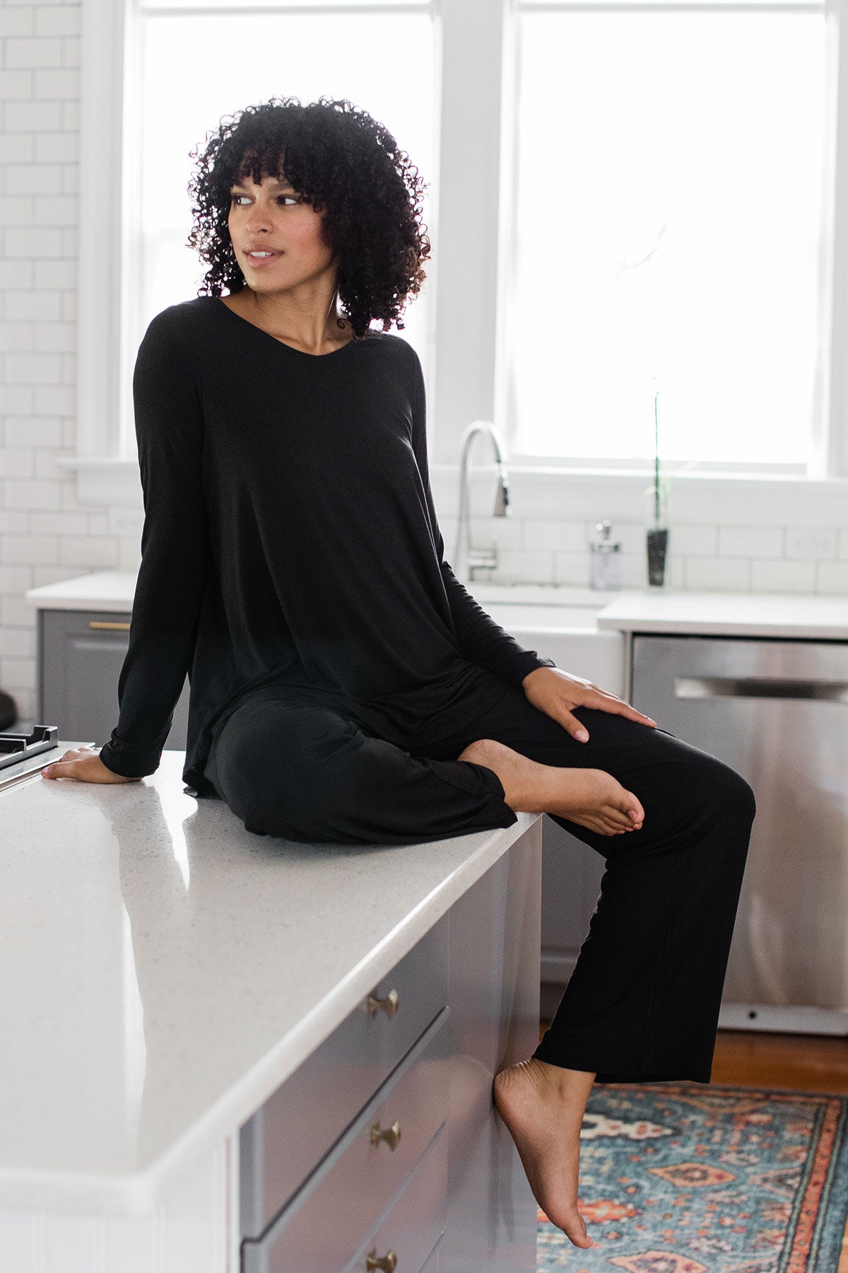 A woman sitting on top of a kitchen countertop and looking off to the side, wearing Yala Norah Long Sleeve Bamboo Pajama Set in Black