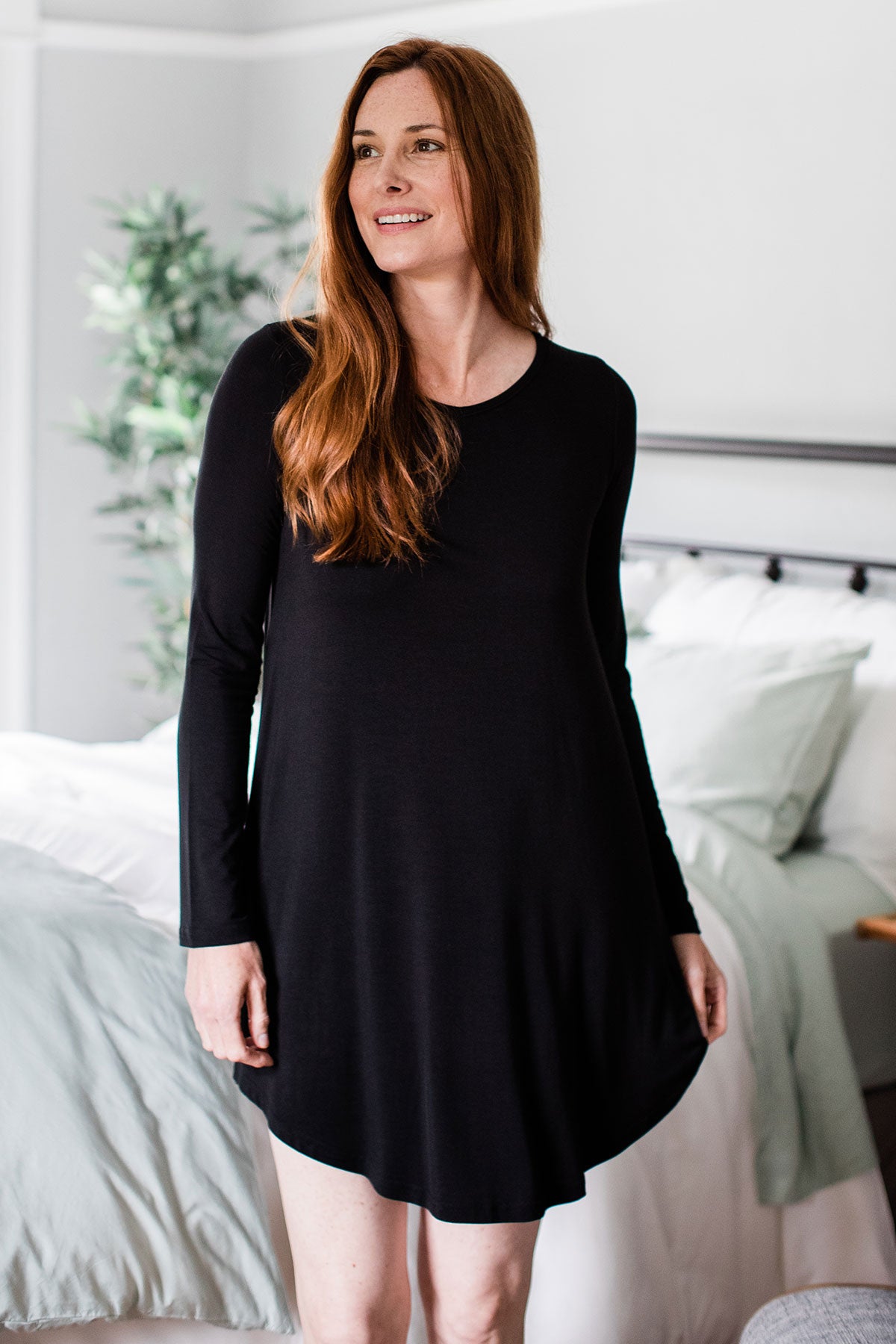 A woman standing and smiling while looking off to the side, wearing Yala Norah Long Sleeve Bamboo Nightshirt in Black