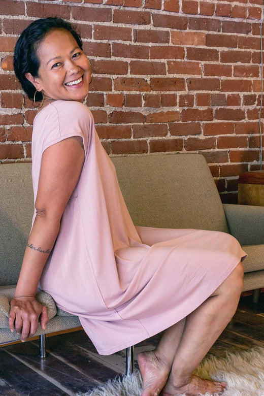 A woman sitting on a couch facing away from the camera and looking back over her shoulder, wearing Yala Opal Swing Bamboo Nightshirt in Lotus Pink
