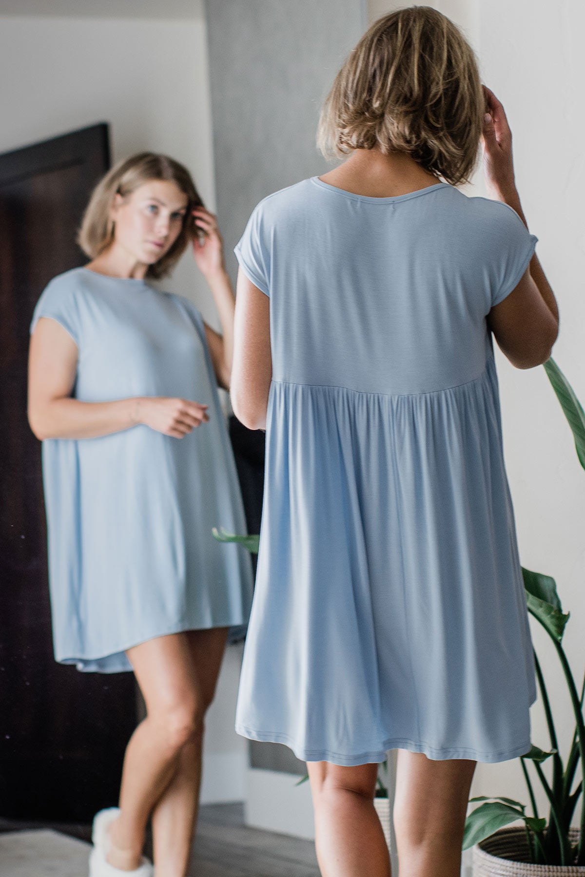A woman standing facing away from the camera in front of a mirror with one hand in her hair, wearing Yala Opal Swing Bamboo Nightshirt in Sky