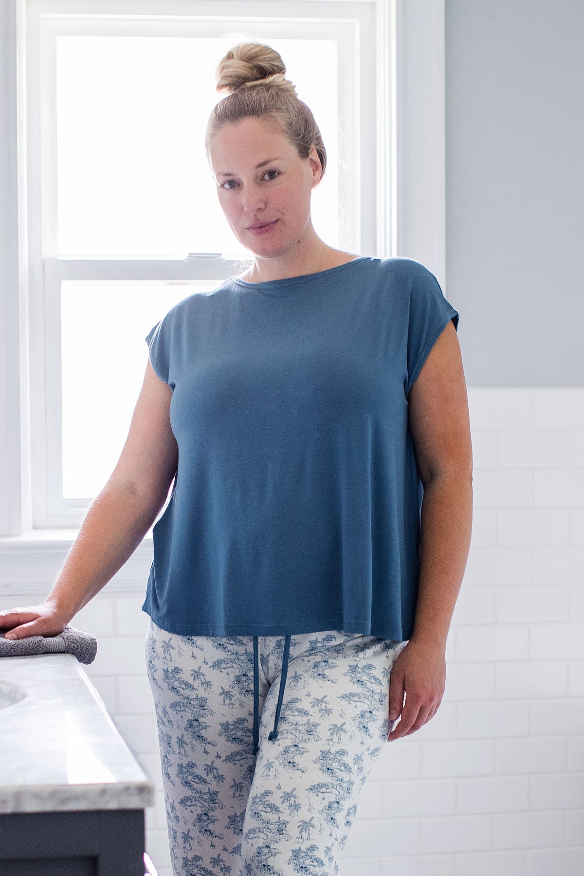 A woman standing with one hand on a countertop, wearing Yala Opal Swing Bamboo Top in Denim