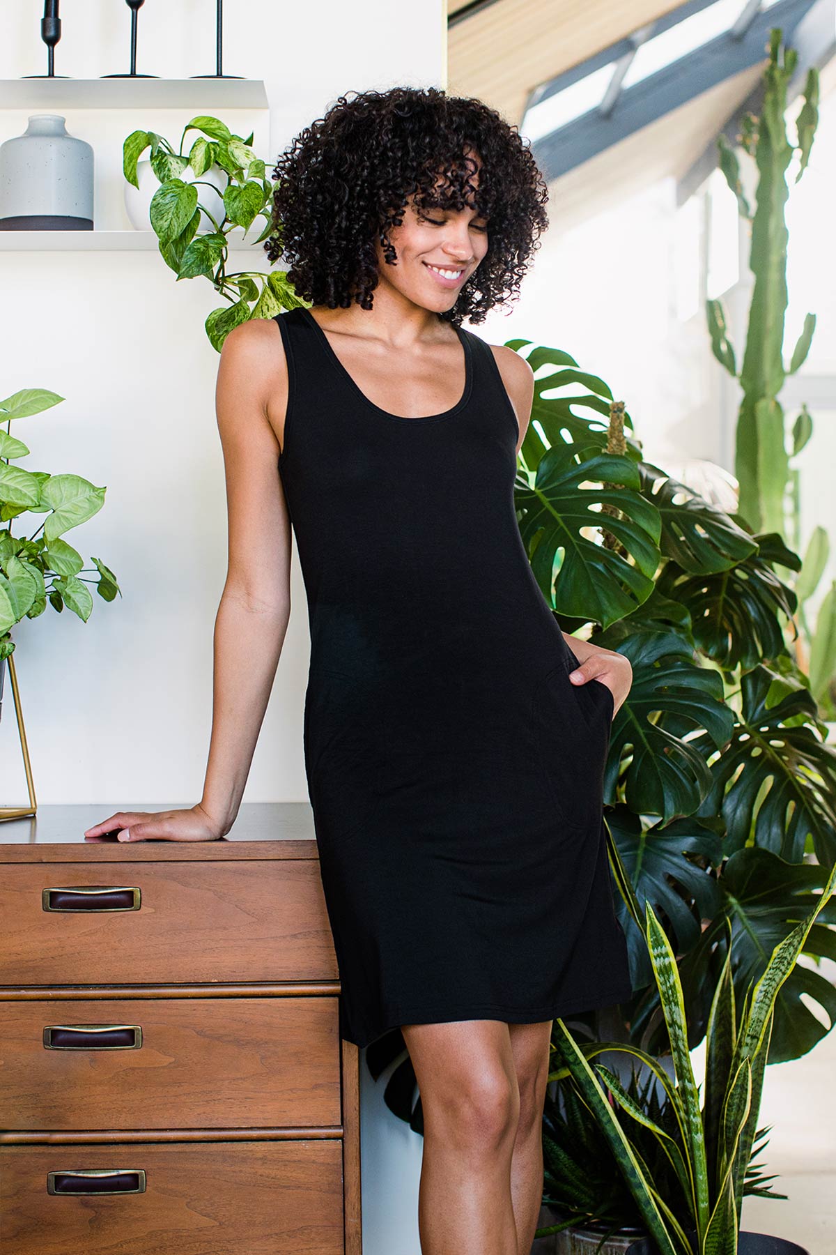 A woman standing and leaning on a dresser with one arm, wearing Yala Riley Racerback Two Pocket Bamboo Shift Dress in Black