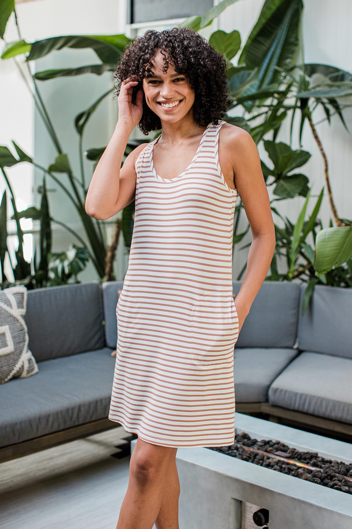 A womand standing andn smiling with one hand in her pocket, wearing Yala Riley Racerback Two Pocket Bamboo Shift dress in Camel Newport Stripe