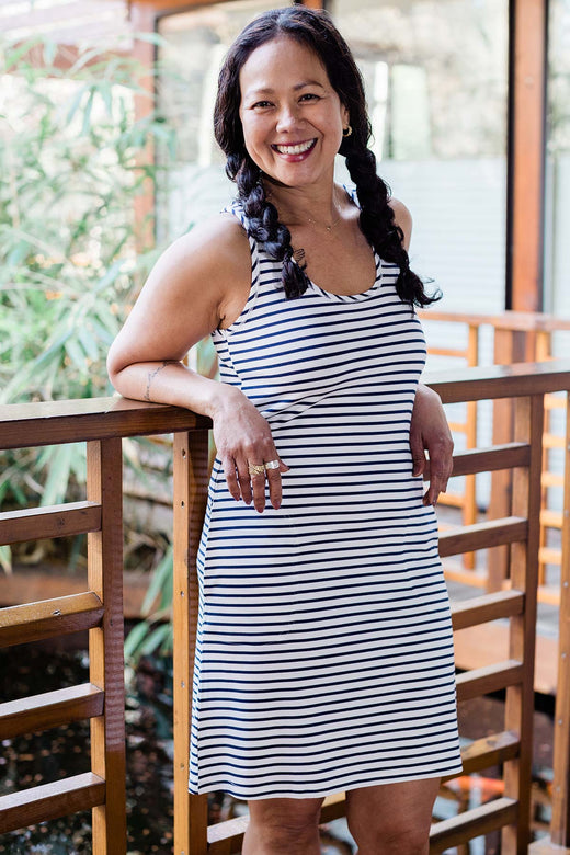 A woman standing and smiling while leaning back with her arms resting on railing, wearing Yala Riley Racerback Two Pocket Bamboo Shift Dress in Navy Newport Stripe