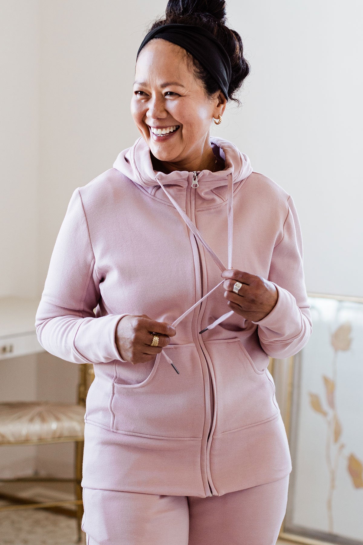 A woman standing and smiling while looking to the side, wearing Yala Scarlet Zip-Up Bamboo and Organic Cotton Hooded Jacket Sweatshirt in Lotus Pink