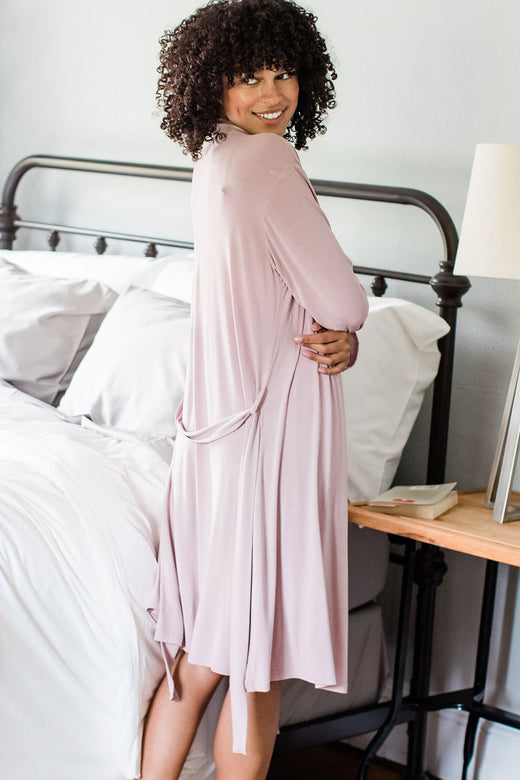 A woman facing away from the camera and looking back over her shoulder with her arms wrapped around herself, wearing Yala Serenity Long Sleeve Short Belted Bamboo Robe in Lotus Pink