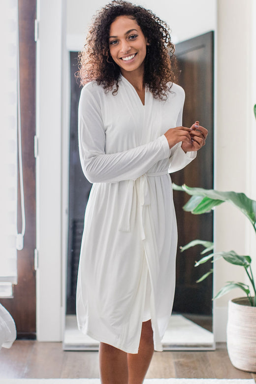 A woman standing and smiling with both hands held to one side, wearing Yala Serenity Long Sleeve Short Belted Bamboo Robe in Natural