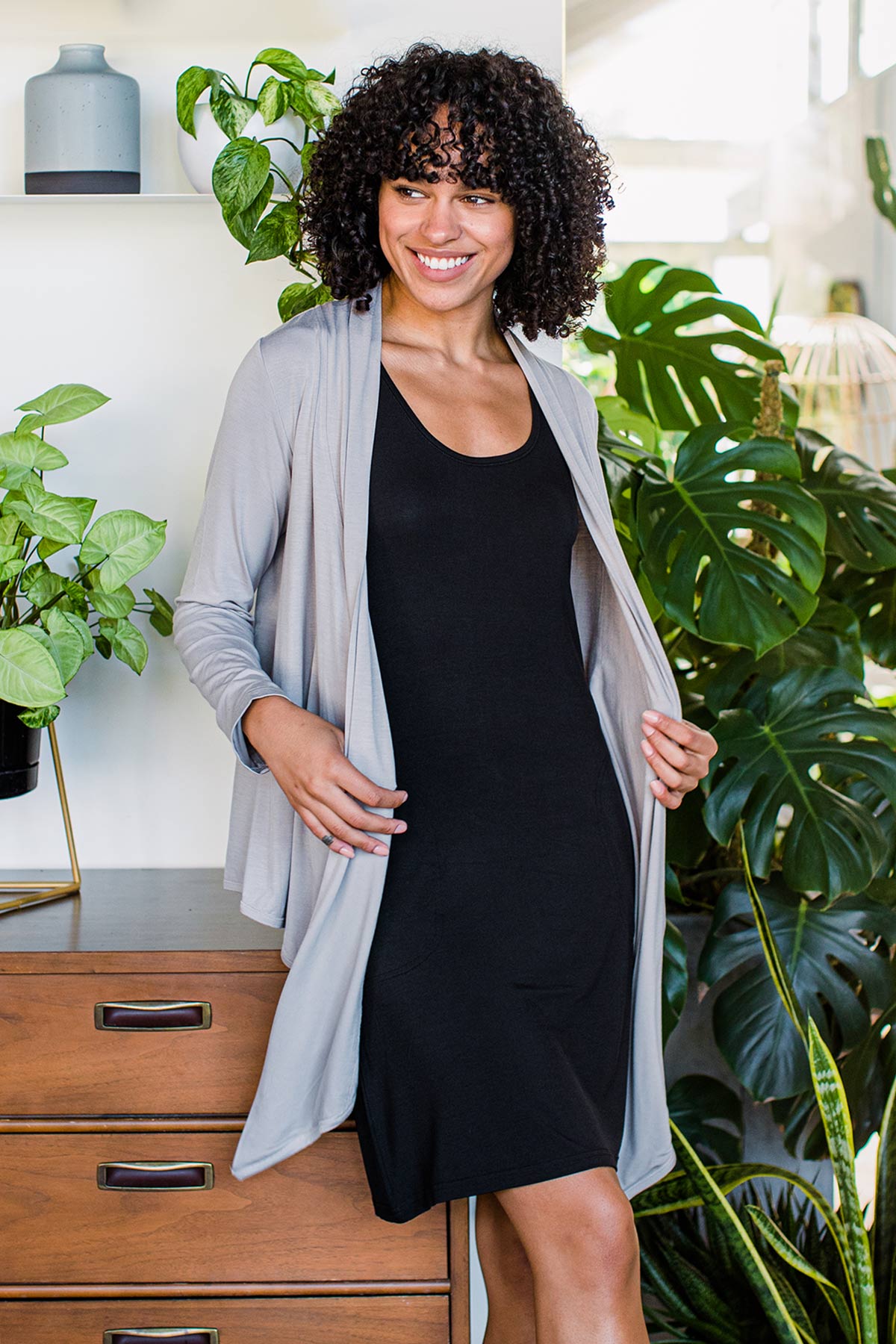 A woman standing and smiling while leaning back against a dresser, wearing Yala Sophie Bamboo Cardigan Wrap in Ash