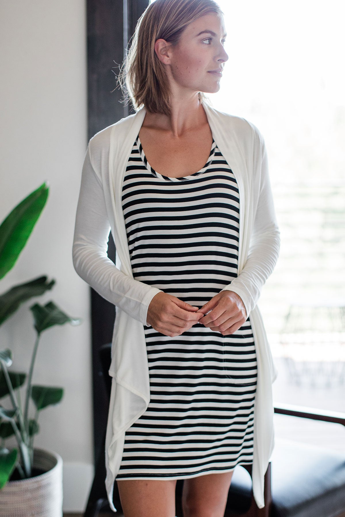 A woman standing and looking off to the side with both hands held in front of her waist, wearing Yala Sophie Bamboo Cardigan Wrap in Natural